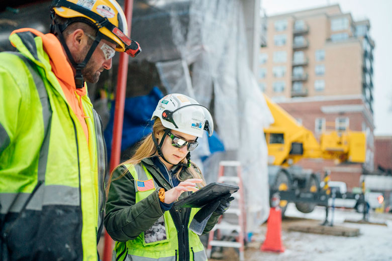 A long haired woman looking at an ipad with a man on a construction site wearing safety helmet and neon vest